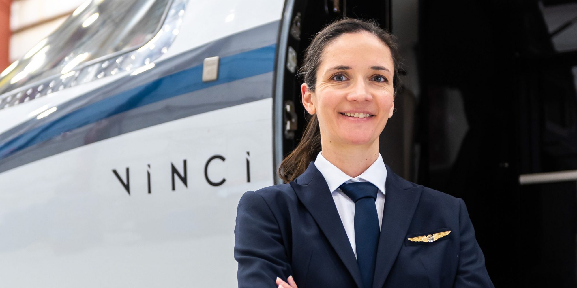 A confident female pilot stands with arms crossed beside a sleek aircraft, wearing a navy uniform and a pilot's wings pin.