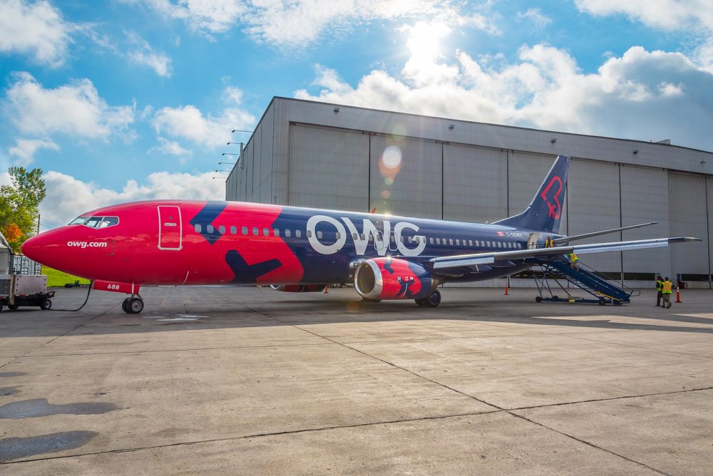 Un avion de ligne aux couleurs vives, rouge et bleu, est stationné devant un hangar. Une échelle est positionnée pour l'embarquement.