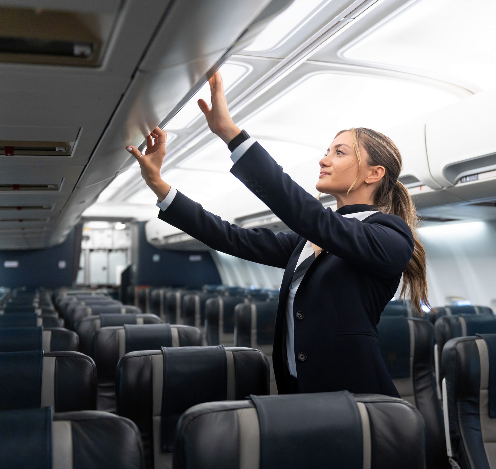 A flight attendant in a navy uniform adjusts the overhead compartment in a nearly empty airplane cabin.