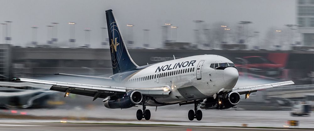 A Noliner aircraft lands on a wet runway, with rain and mist creating a dramatic backdrop at an airport.