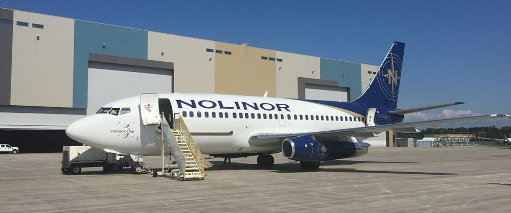 Un avion de la compagnie Nolinor est stationné sur le tarmac, avec une échelle d'embarquement à côté. Des bâtiments en arrière-plan.