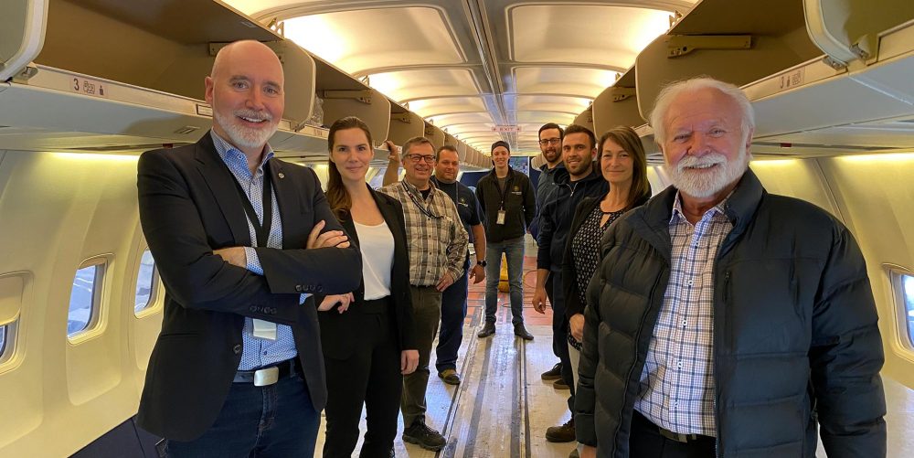 Un groupe de neuf personnes souriantes se tient dans un couloir d'avion, avec des sièges visibles de chaque côté.