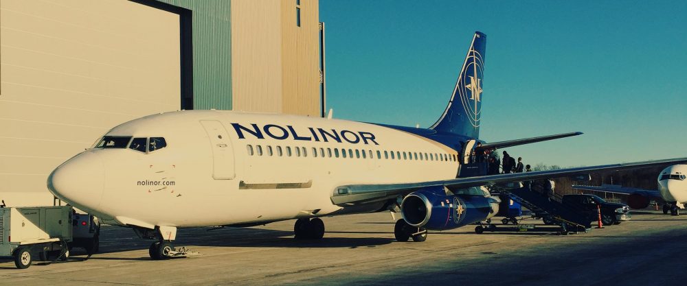 Un avion de la compagnie Nolinor est stationné sur le tarmac, des passagers montent à bord par une échelle. Le ciel est dégagé.