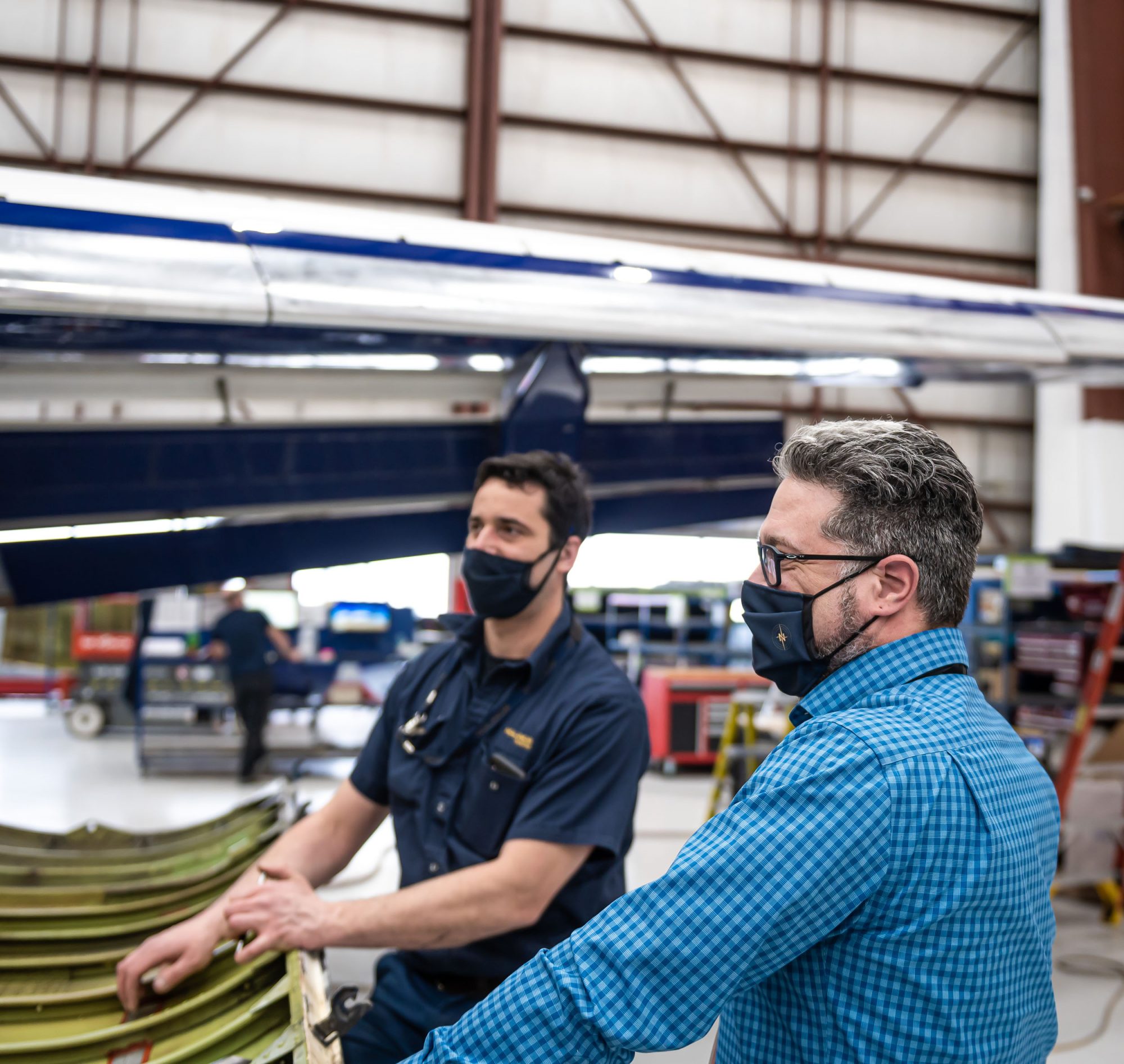 Two technicians in masks discuss aircraft components in a hangar, with a large aircraft wing visible in the background.