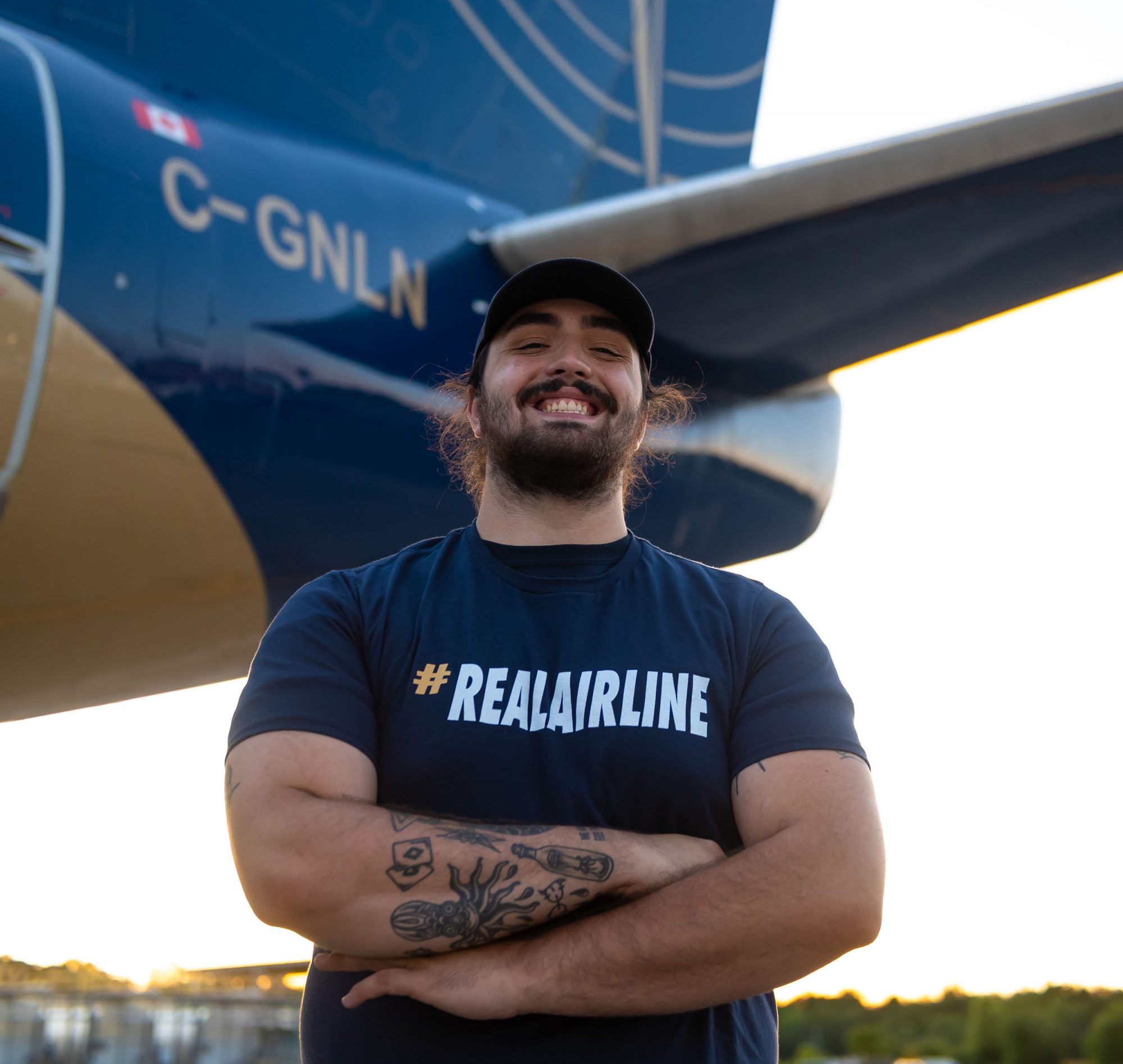 A smiling man stands with arms crossed in front of an aircraft, wearing a "#REALAIRLINE" t-shirt. The setting is outdoors during sunset.