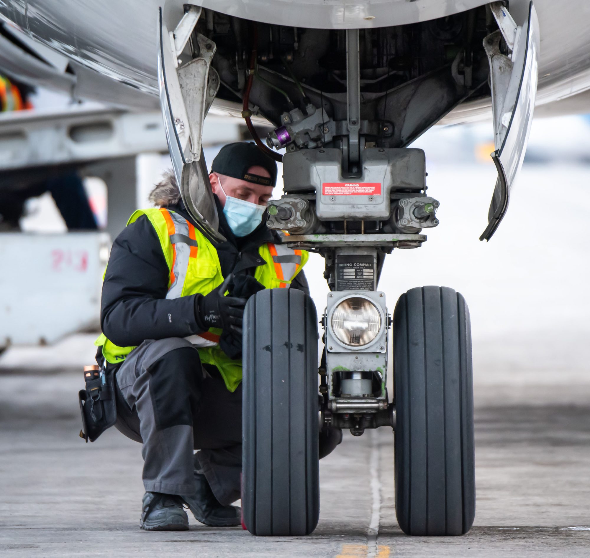 A technician in a safety vest inspects the landing gear of an aircraft on the tarmac, wearing a mask for safety.