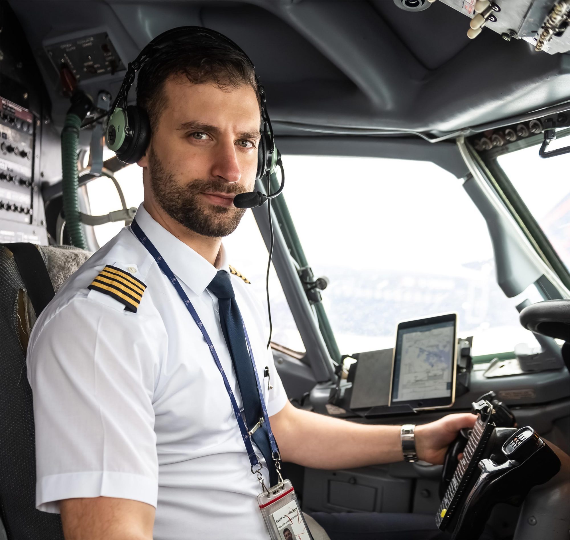 Pilot in the cockpit wearing a headset, looking at the camera with flight instruments and a tablet displaying navigation.