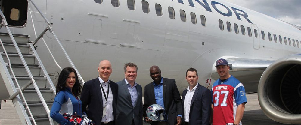 Un groupe de six personnes pose près d'un avion, avec des membres d'une équipe de football en tenue. Des pom-pom girls sont présentes, tenant un casque de football.