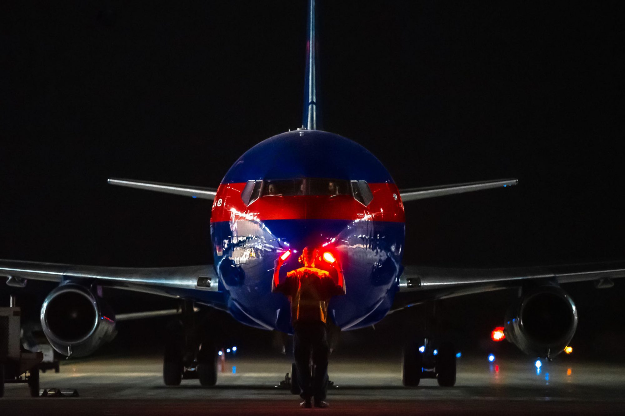 A ground crew member signals to a brightly colored airplane at night, with runway lights illuminating the scene.