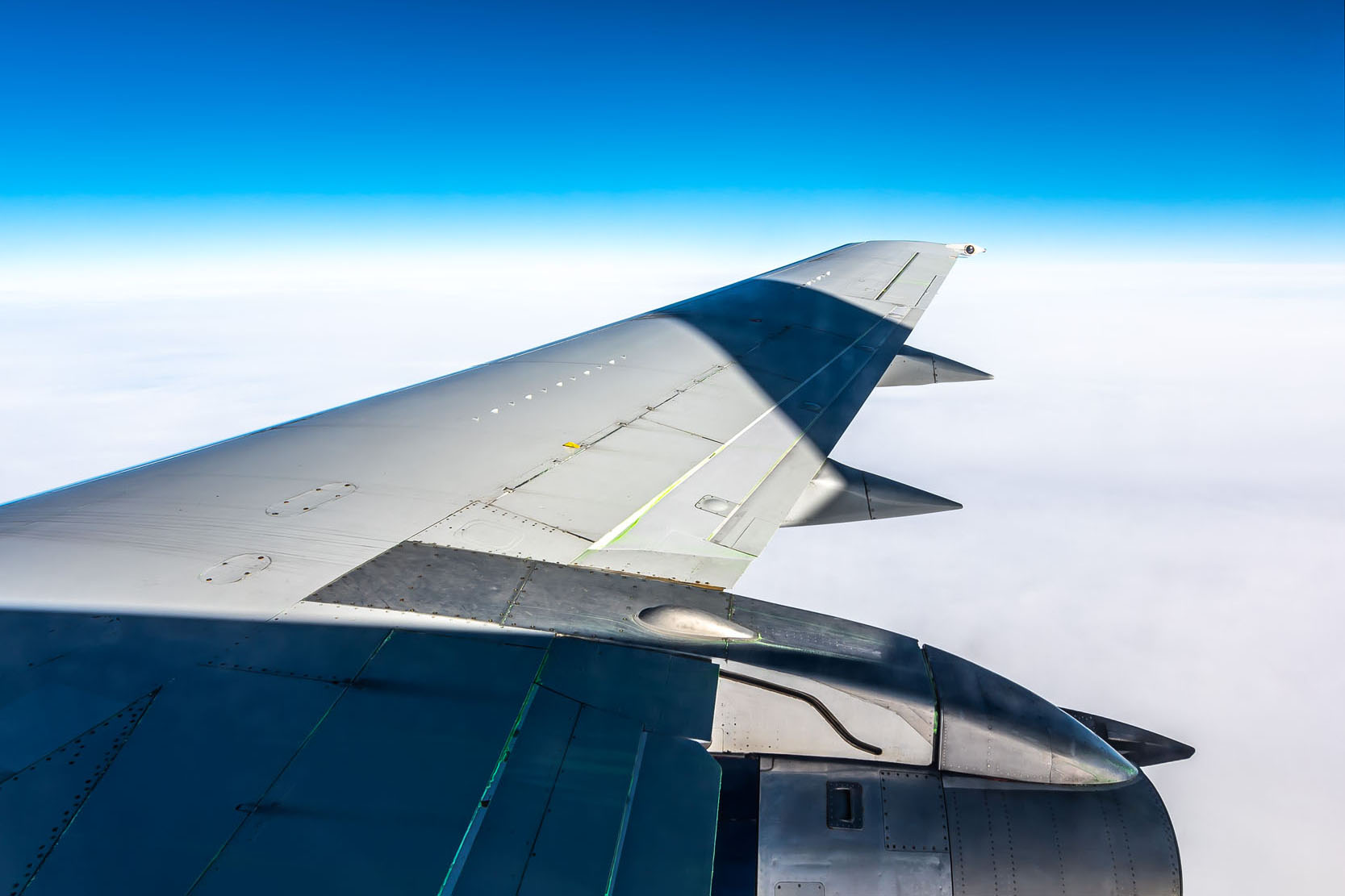 Airplane wing extending into a clear blue sky above a blanket of white clouds, showcasing the wing's details and structure.