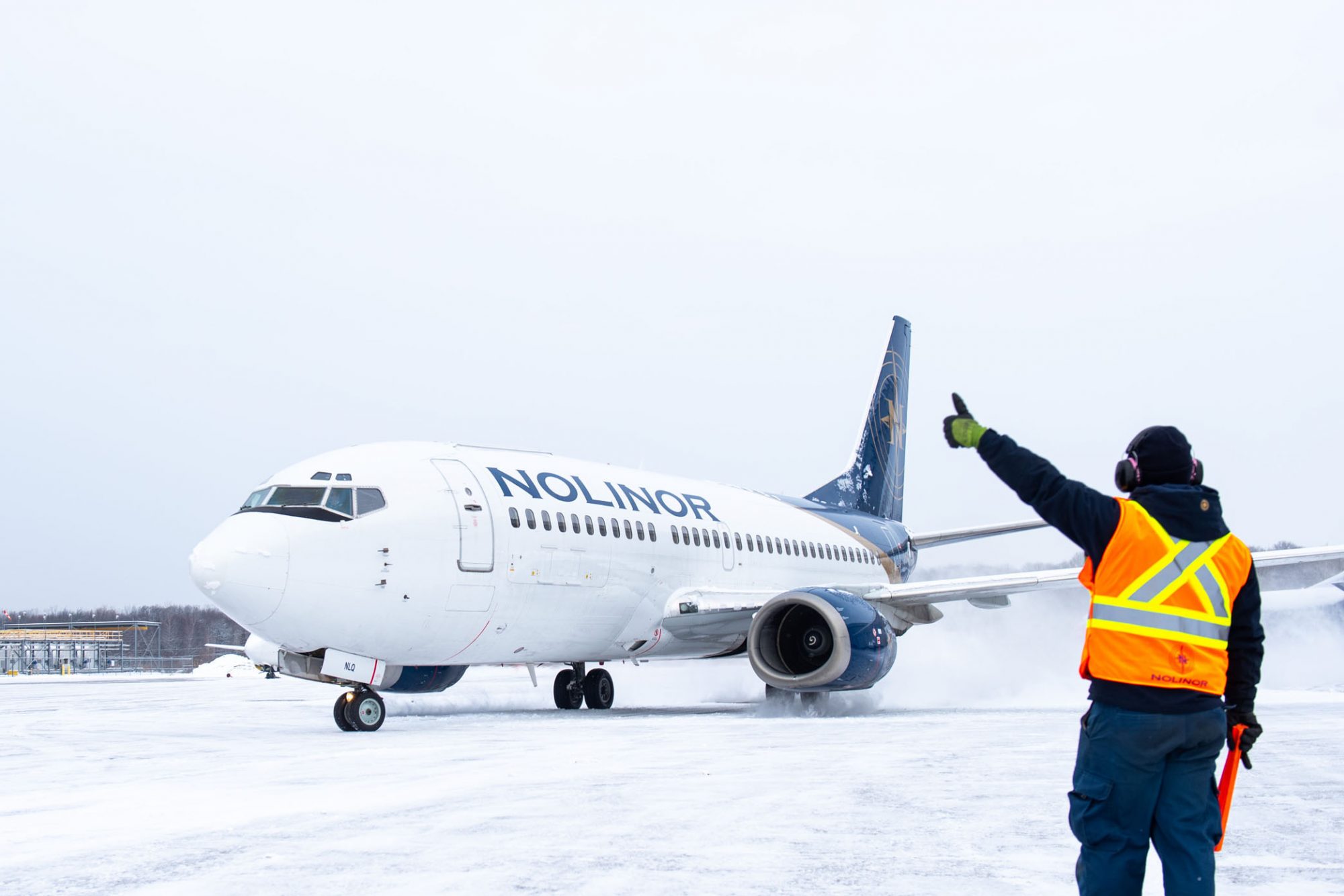 A ground crew member in a safety vest directs a Noliner Airlines plane on a snowy runway.