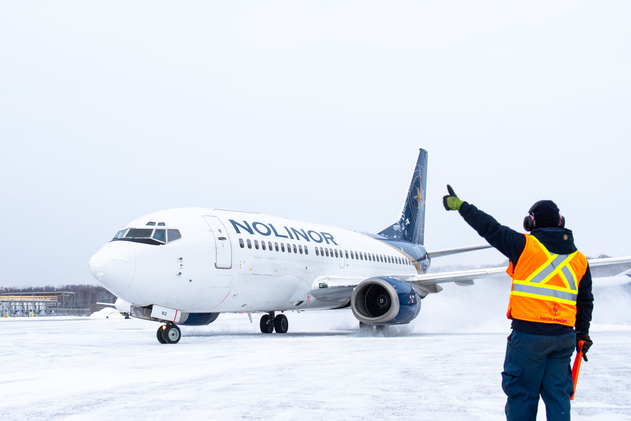 A ground crew member in a safety vest directs a Noliner Airlines plane on a snowy runway.
