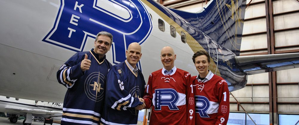 Quatre hommes posent devant un avion, souriant et portant des maillots de hockey. Deux maillots sont rouges avec le logo "Rocket".
