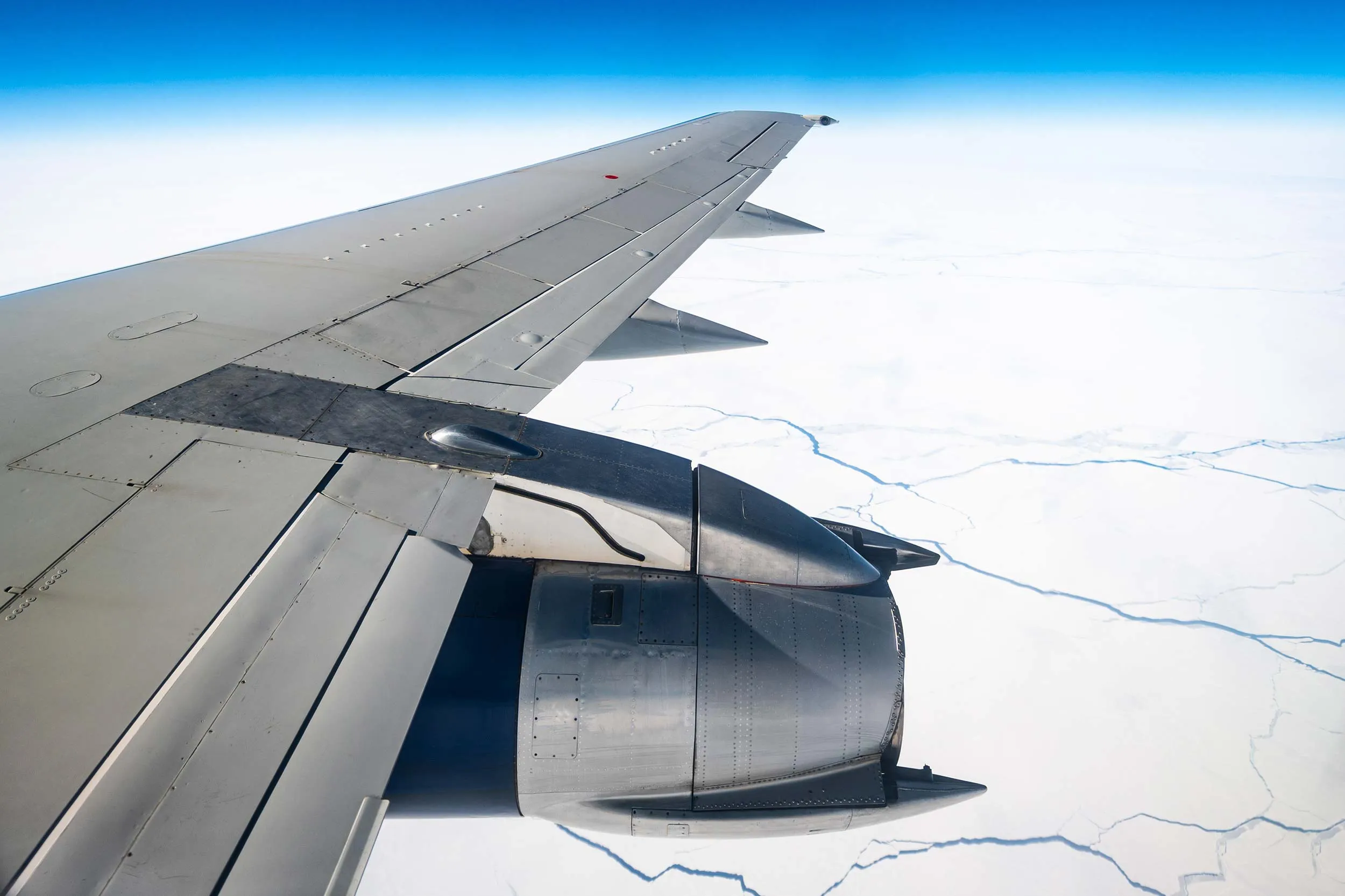 View from an airplane wing showing a snowy landscape below, with a jet engine and wing flap visible against a clear blue sky.