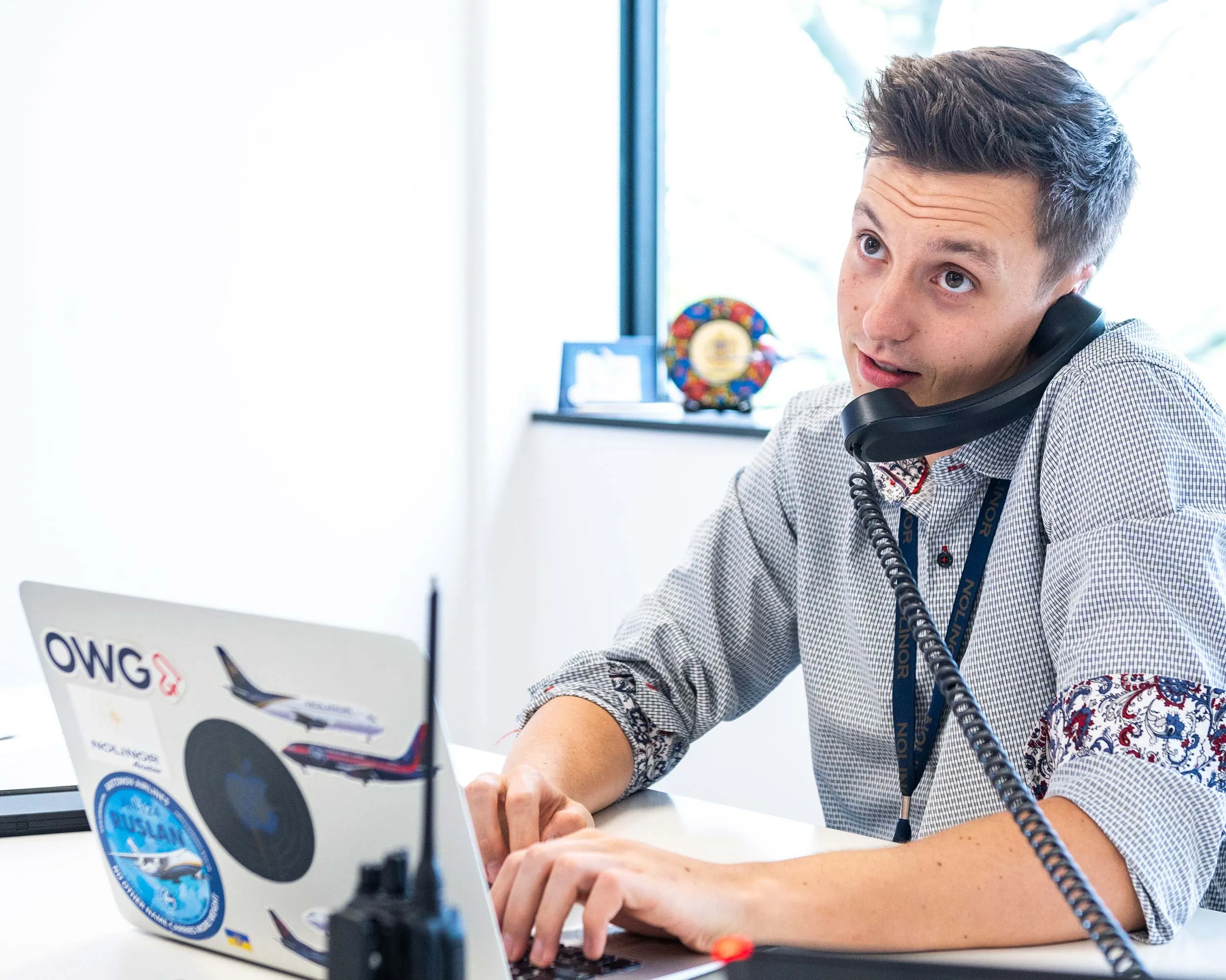 A young man with short hair talks on the phone while typing on a laptop covered in stickers, seated at a desk by a window.