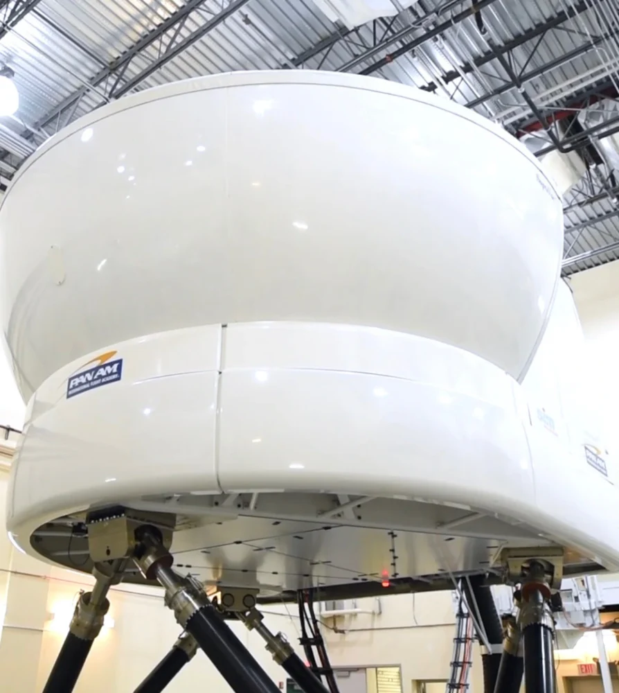 Simulateur de vol blanc installé sur des supports robustes, dans un environnement intérieur. Le logo "PWAM" est visible sur le côté.