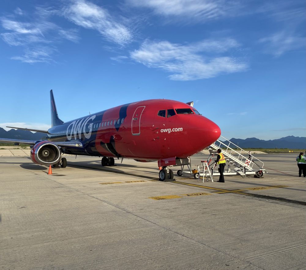 Un avion de la compagnie OWG est stationné sur le tarmac, avec des escaliers d'embarquement et des agents au sol. Le ciel est dégagé.