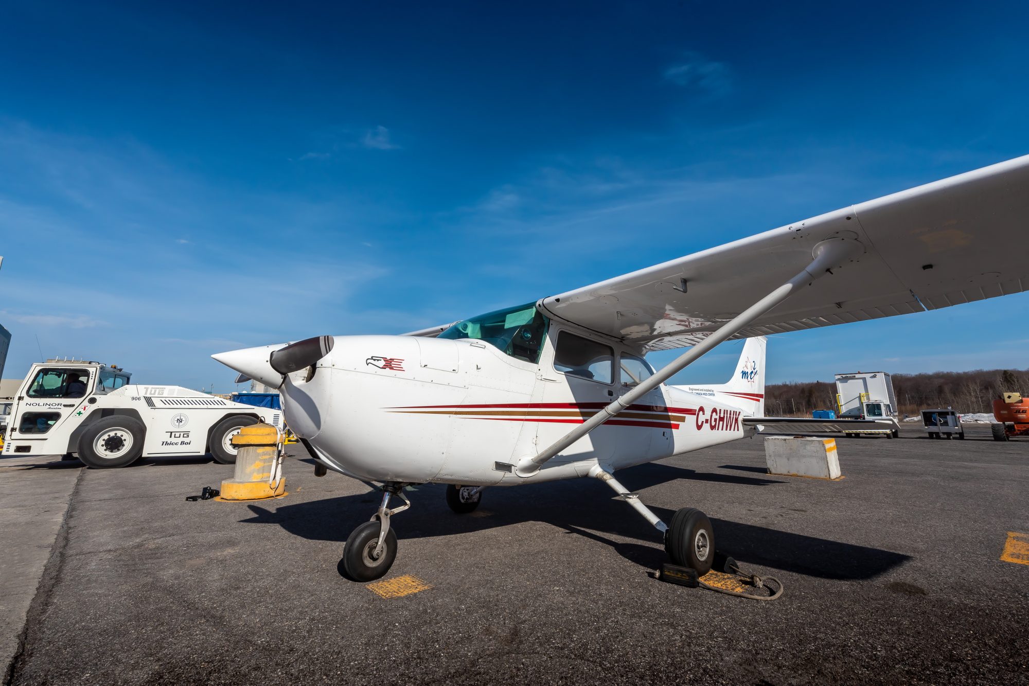 A small Cessna aircraft parked on a tarmac, with a blue sky in the background and ground support equipment nearby.