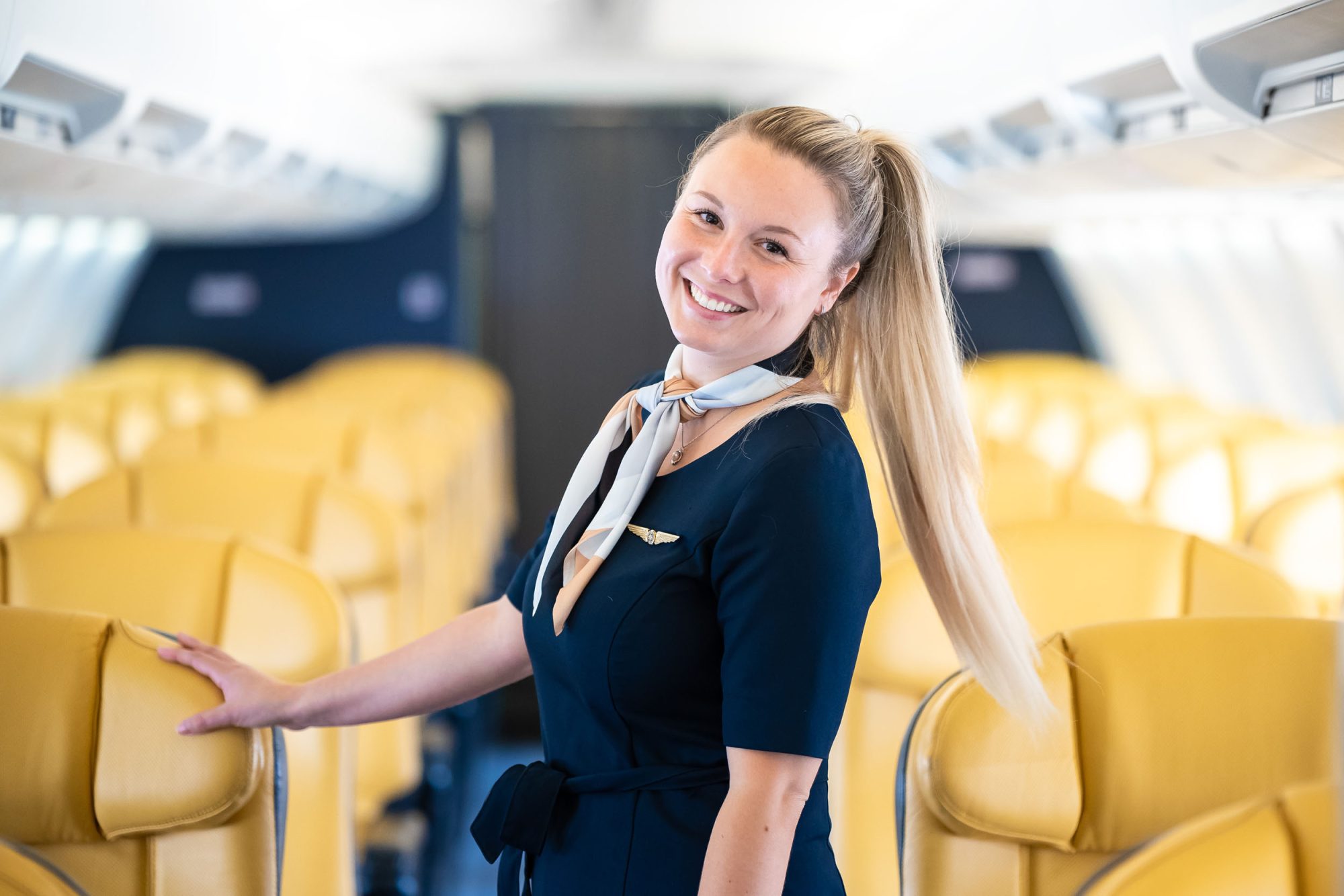 A flight attendant smiles while standing in an airplane aisle, surrounded by yellow seats, ready to assist passengers.