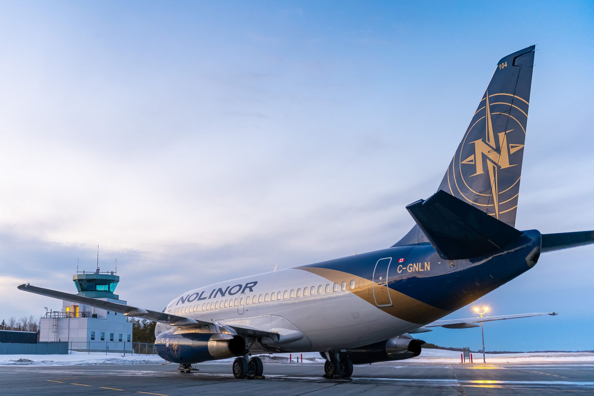 Nolinor Airlines aircraft parked on a tarmac with a control tower in the background during twilight.