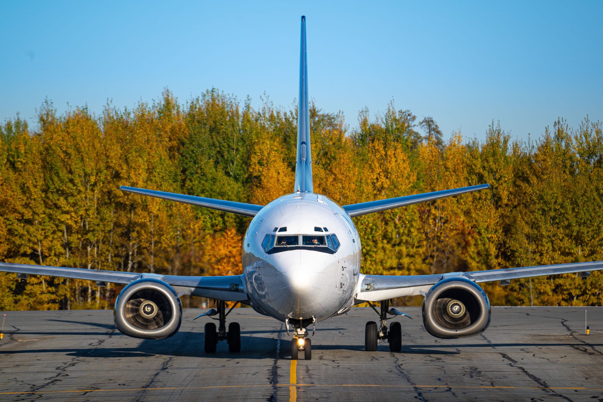 A commercial airplane is taxiing on a runway, with a backdrop of vibrant autumn trees in shades of yellow and orange.