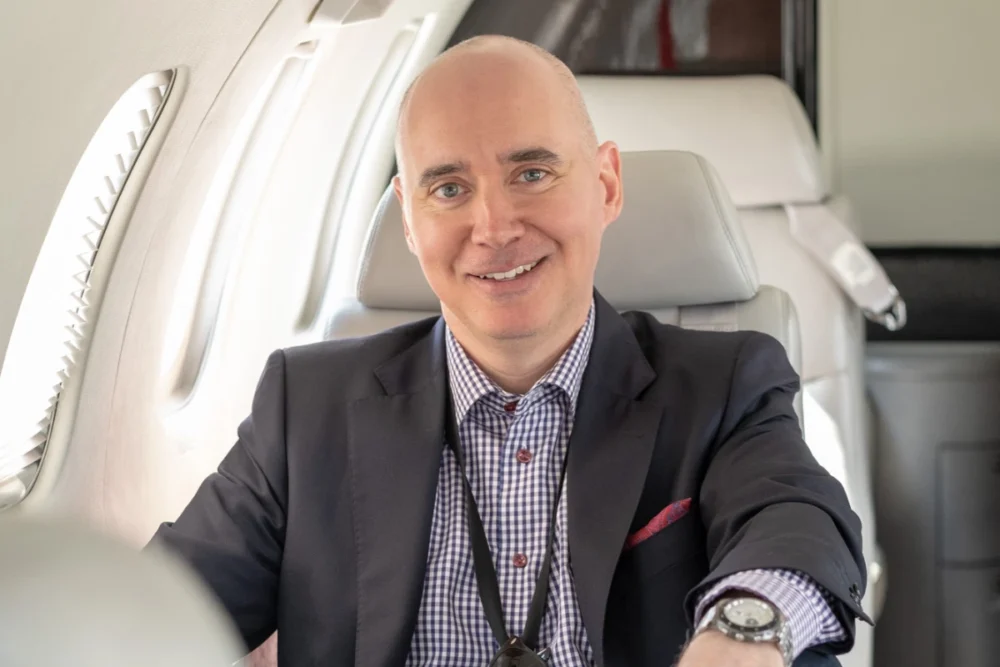 Un homme souriant en costume assis dans un jet privé, avec des sièges en cuir et des fenêtres donnant sur l'extérieur.