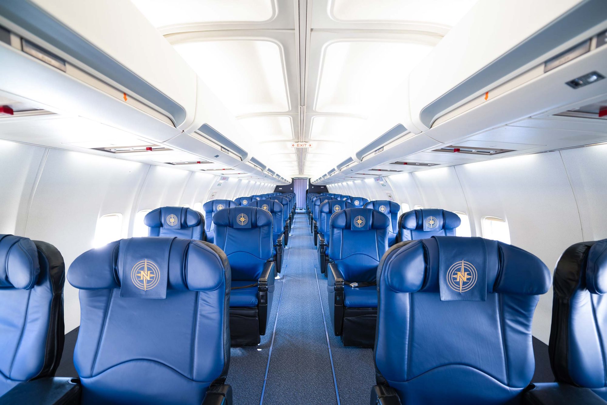 Interior of an airplane featuring rows of blue leather seats with a logo on the headrests, well-lit and spacious aisle.