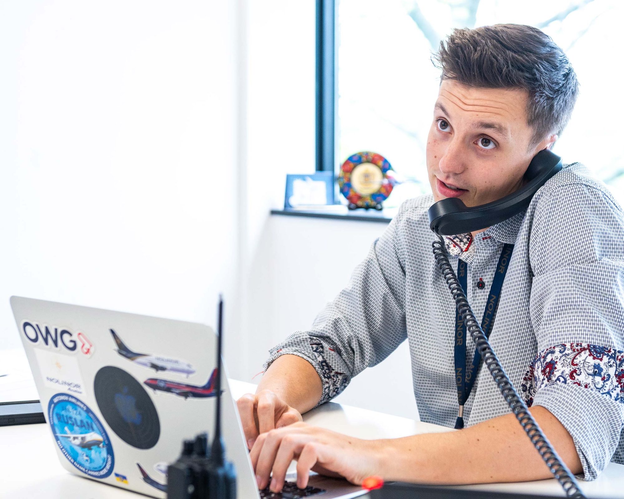 A young man with short hair talks on the phone while typing on a laptop covered in stickers, seated at a desk by a window.