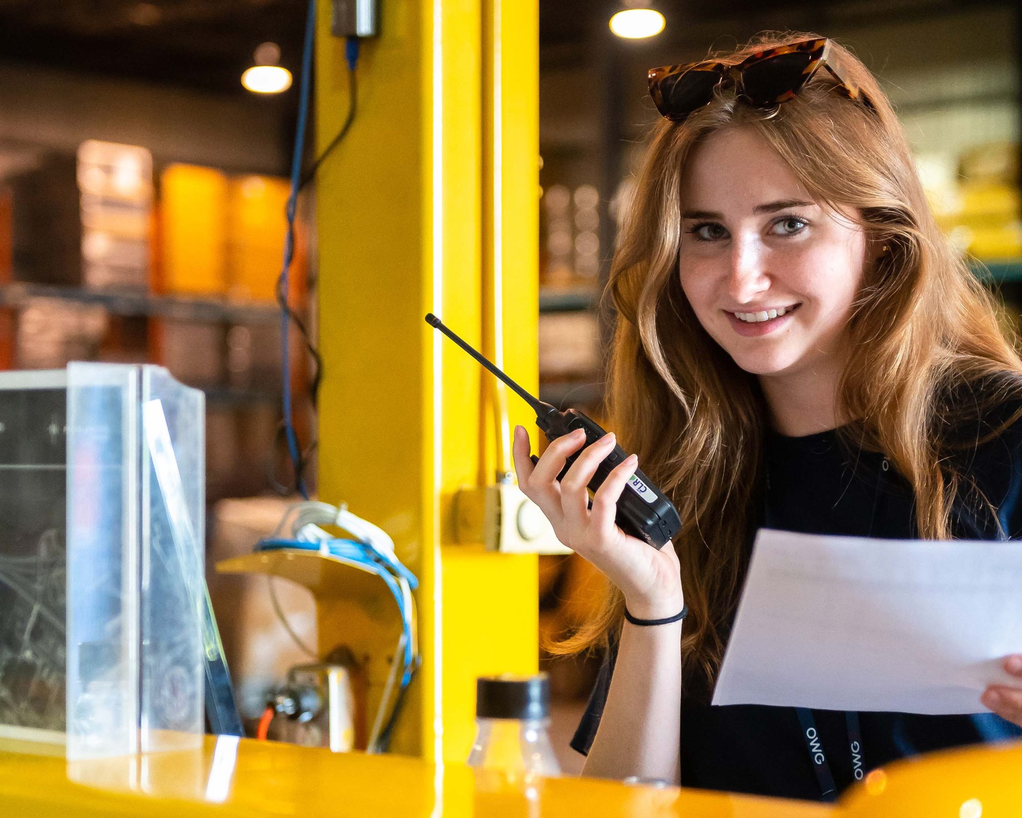 A young woman with long hair holds a walkie-talkie and a paper while smiling in a warehouse setting.