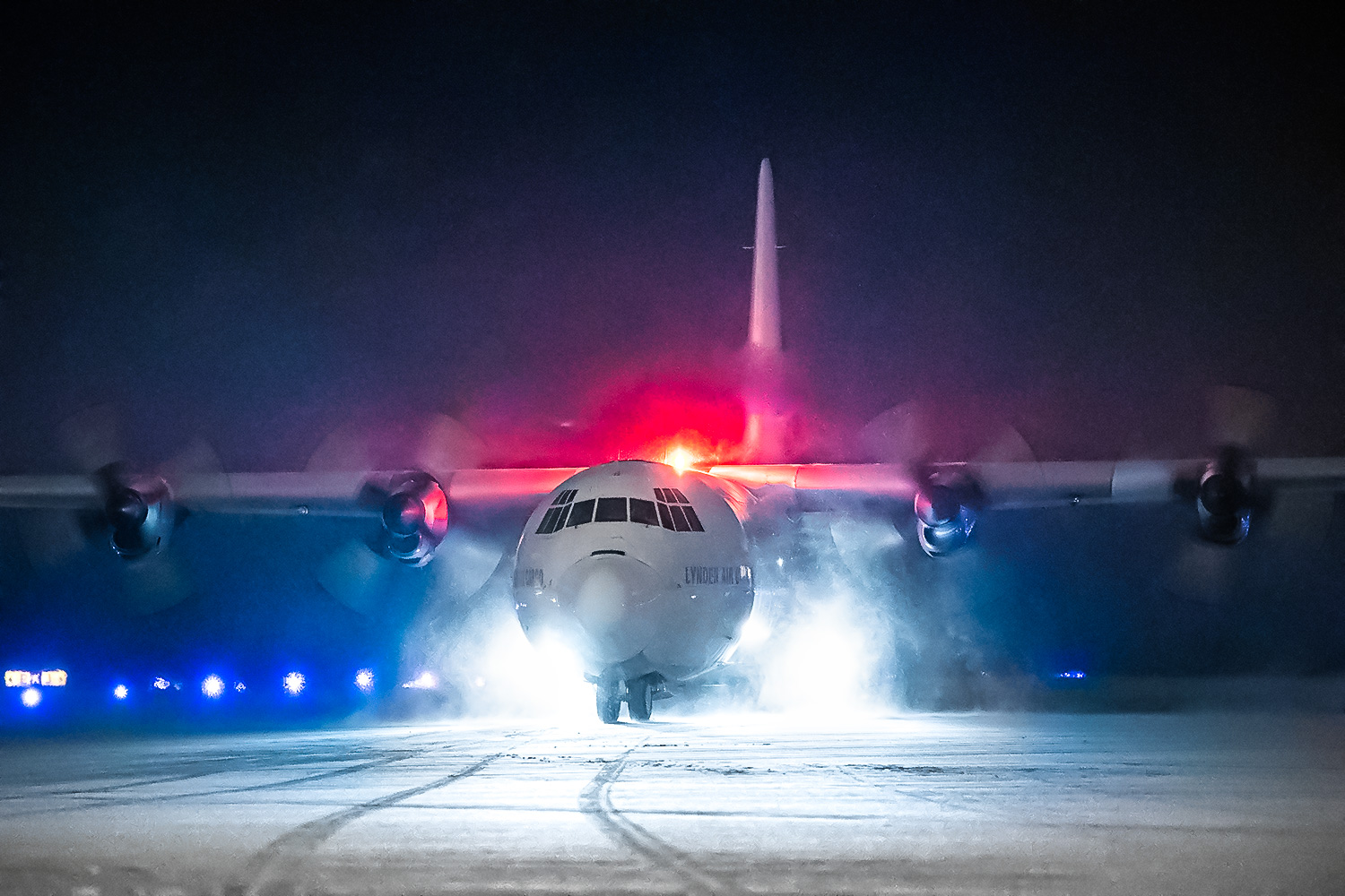 A military aircraft with lights flashing taxis on a snowy runway at night, surrounded by a misty atmosphere.