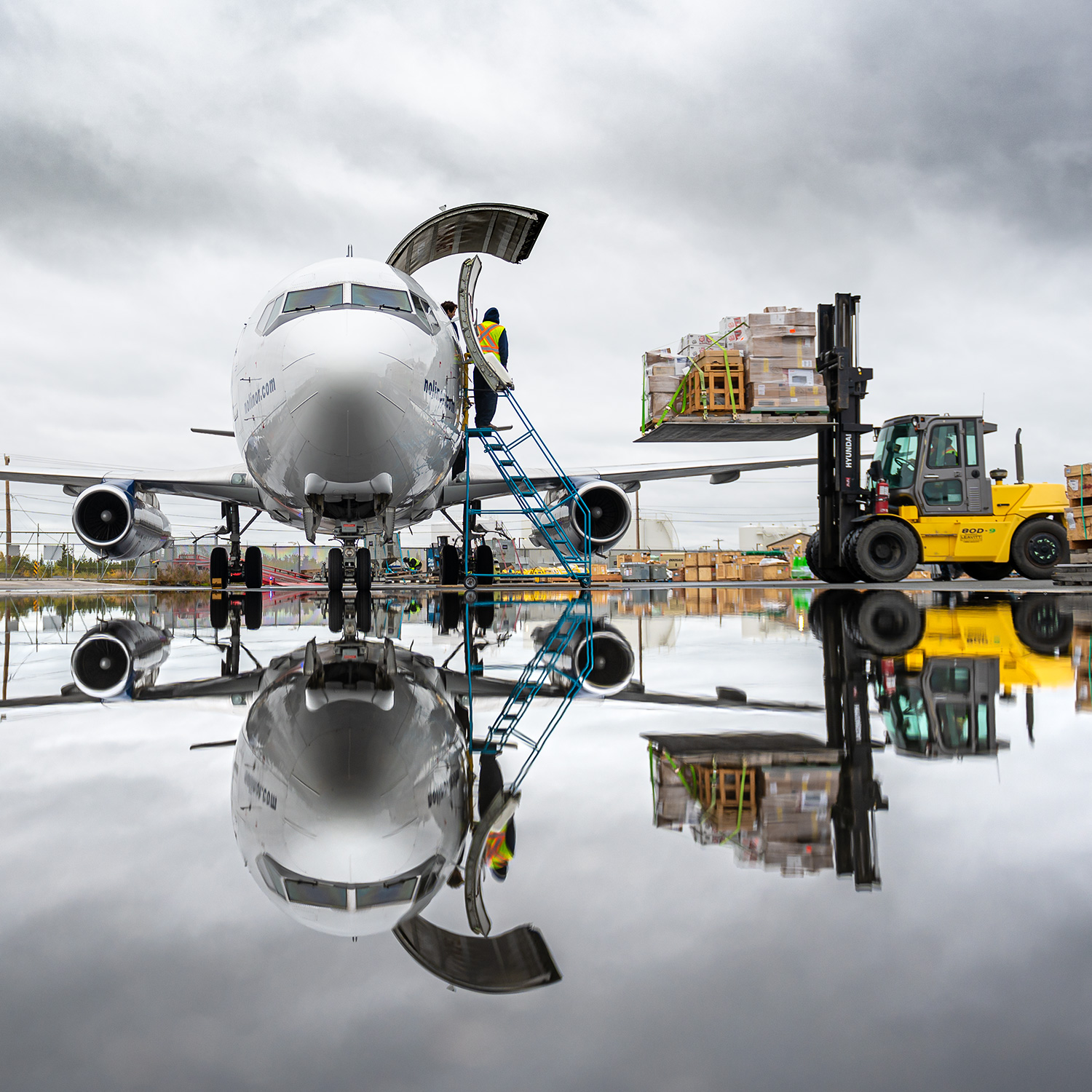 A cargo plane is being loaded with boxes by a forklift, reflecting in a wet surface under a cloudy sky.