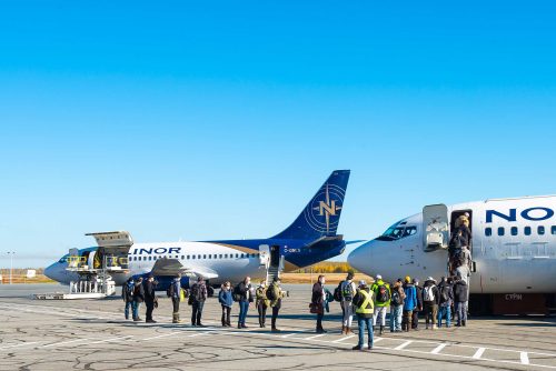 Passengers disembark from a plane while others wait near two parked aircraft under a clear blue sky.