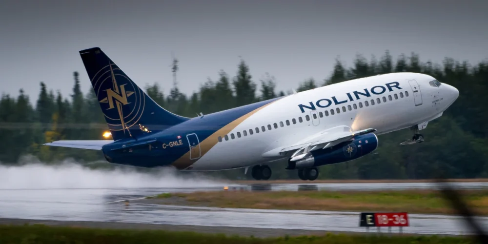 Un avion de la compagnie Nolinor décolle sur une piste humide, soulevant des éclaboussures d'eau sous ses roues. Le ciel est nuageux.