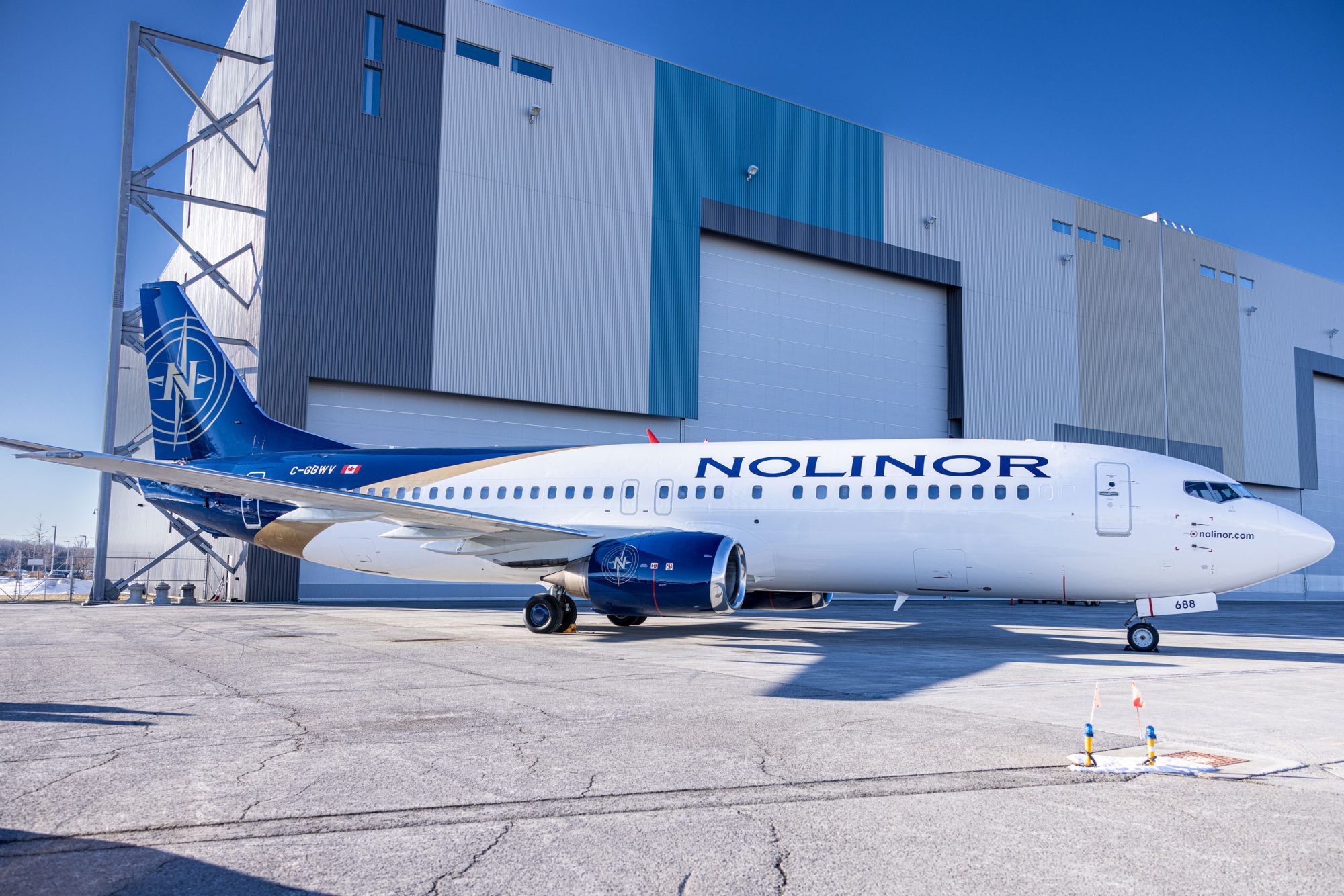 Un avion de la compagnie Nolinor est stationné sur le tarmac devant un hangar moderne, sous un ciel bleu clair.