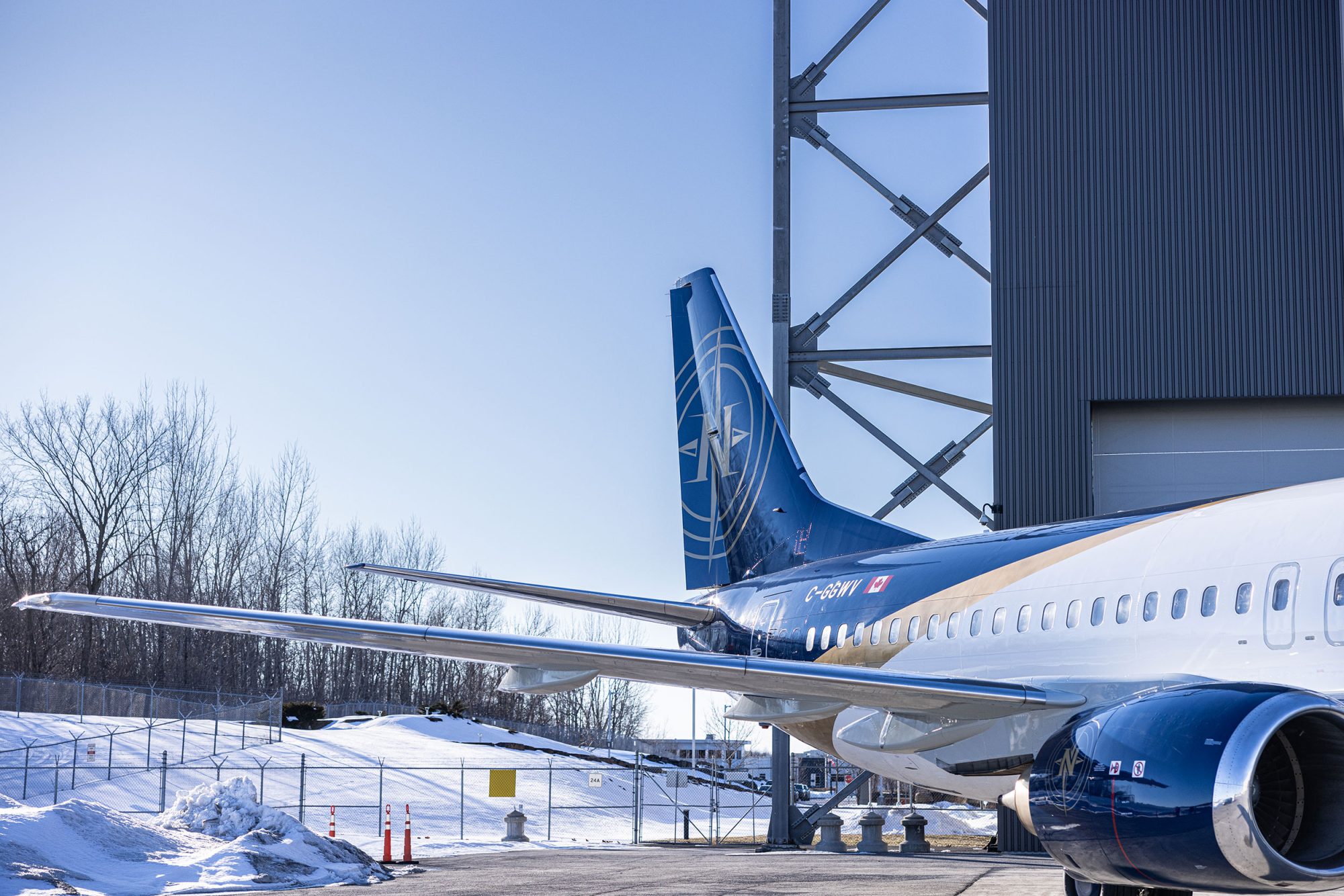 Un avion de ligne stationné près d'un bâtiment, avec un ciel bleu clair en arrière-plan et de la neige au sol. Les ailes et la queue de l'avion sont bien visibles.