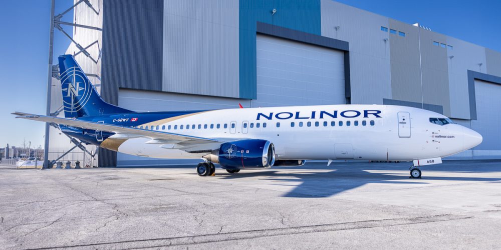 Un avion de la compagnie Nolinor est garé devant un grand bâtiment industriel, avec un ciel dégagé en arrière-plan.