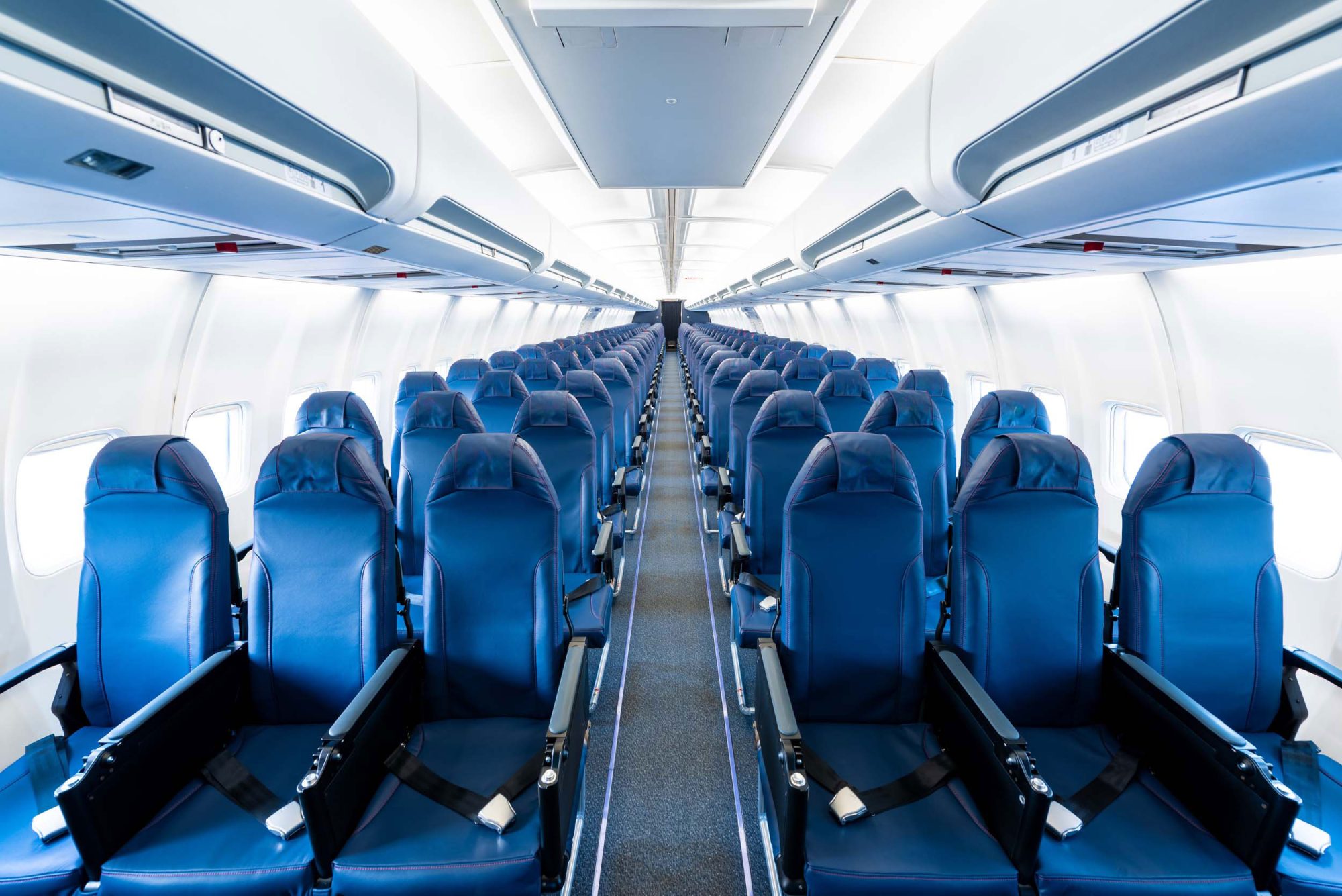 Cabine d'avion avec rangées de sièges bleus vides, lumière naturelle entrant par les fenêtres, allée centrale dégagée.
