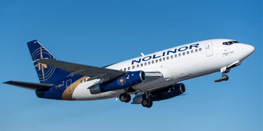 Un avion de la compagnie Nolinor décolle dans un ciel bleu dégagé, montrant son fuselage blanc et ses ailes grises.