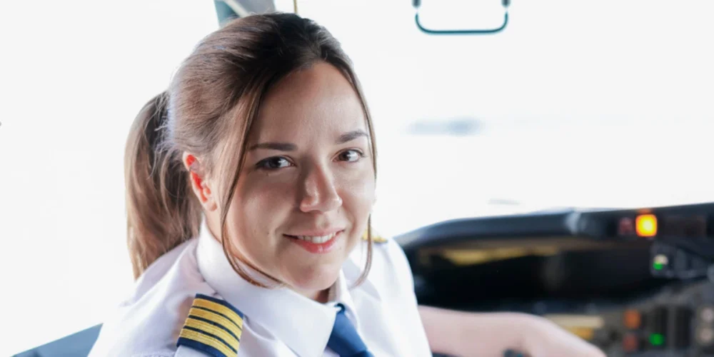 Une pilote souriante est assise dans le cockpit, portant une chemise blanche et des épaulettes dorées. Des instruments de vol sont visibles en arrière-plan.