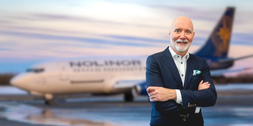 Un homme souriant en costume se tient devant un avion de la compagnie Nolior, sur fond de ciel coloré au crépuscule.