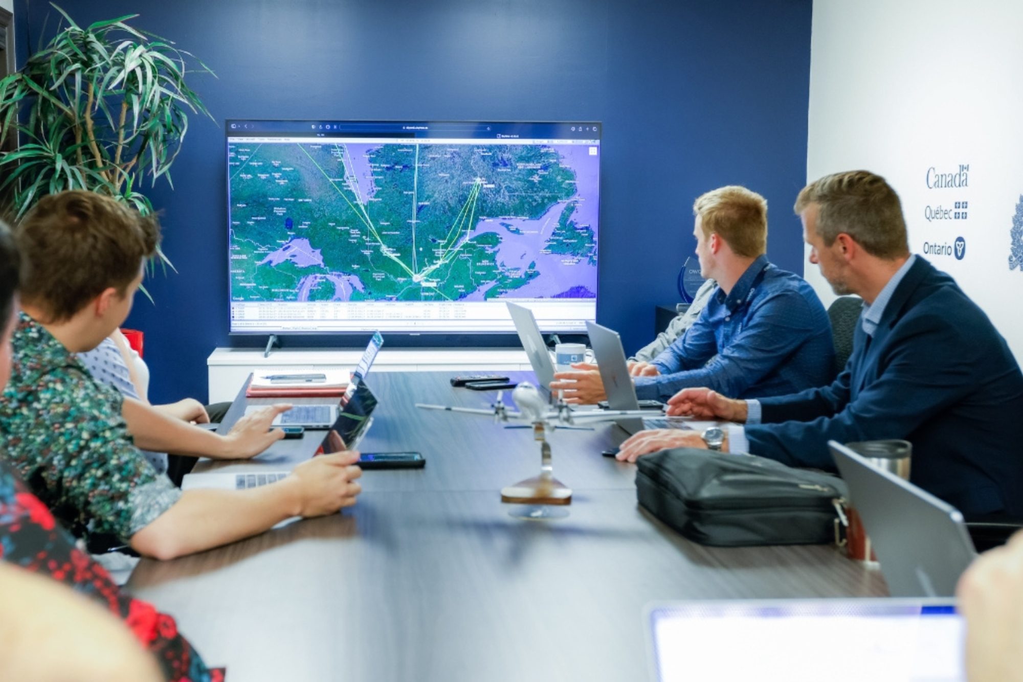 A group of professionals in a meeting room observes a large screen displaying a map with flight paths and data.