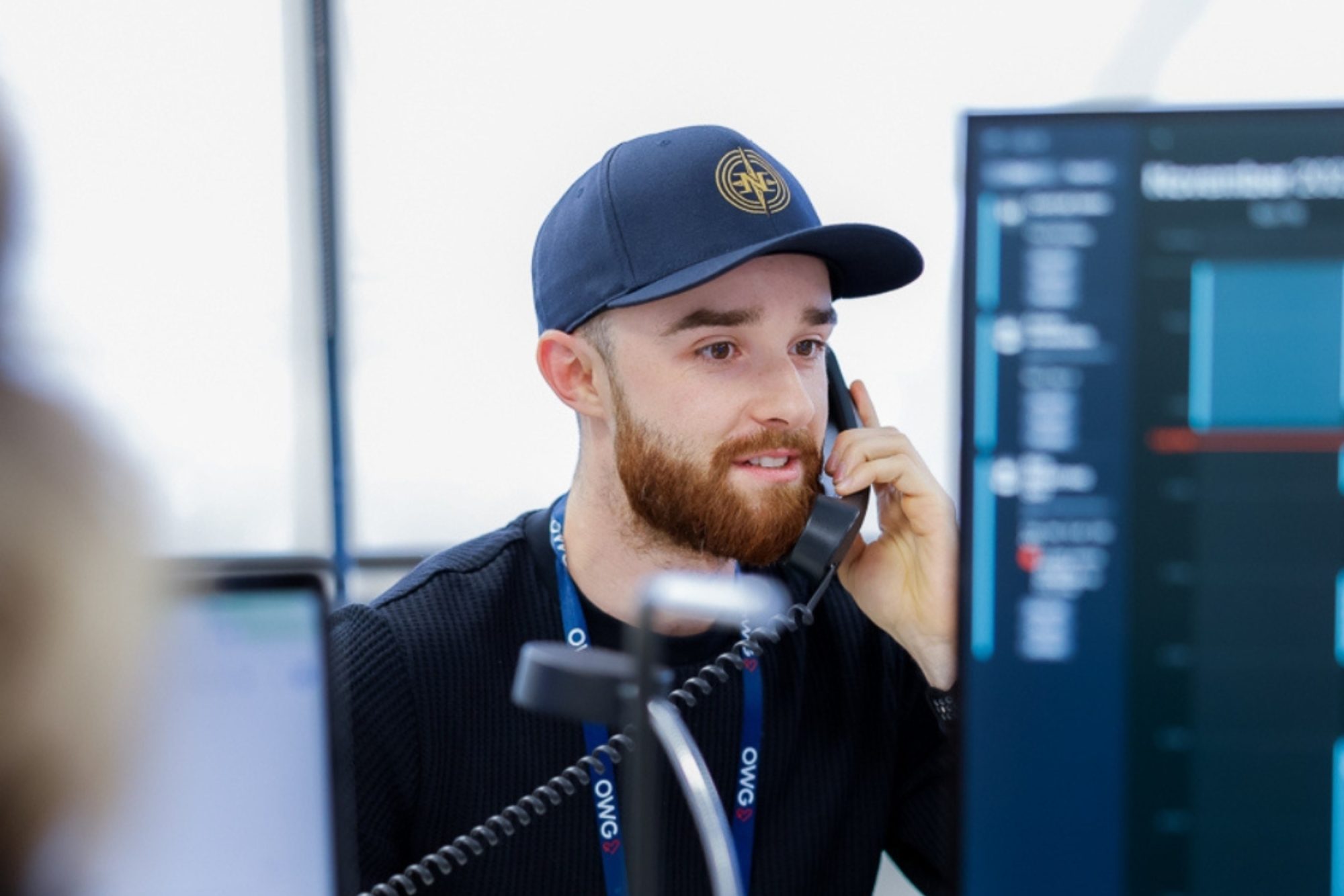 A man with a beard wearing a cap speaks on the phone, focused on a computer screen with data charts in a modern office setting.