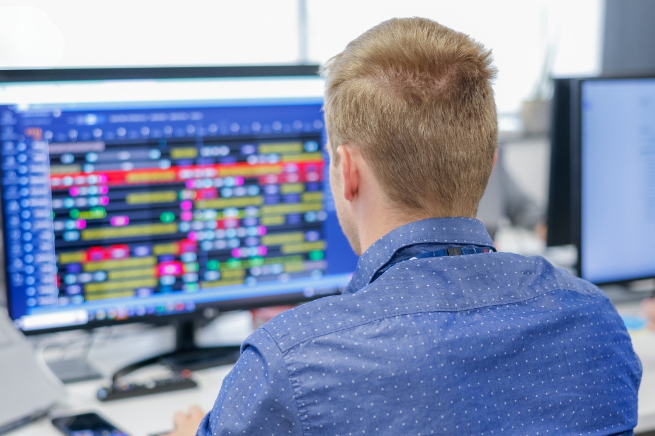 A person with short blonde hair sits at a desk, focused on multiple computer screens displaying colorful data and charts.
