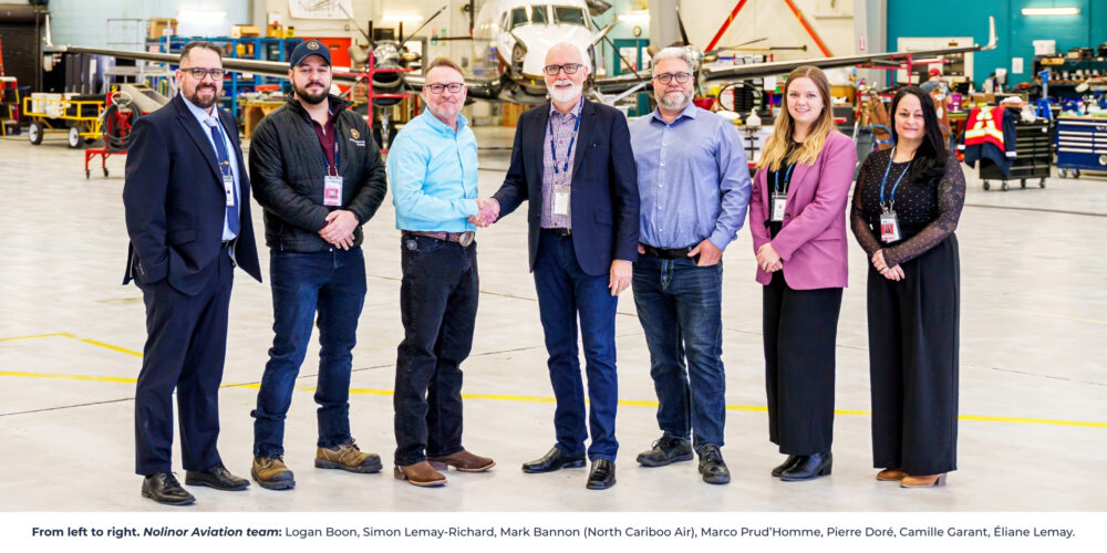A group of seven professionals stands in an aircraft hangar, smiling and shaking hands, with a plane in the background.