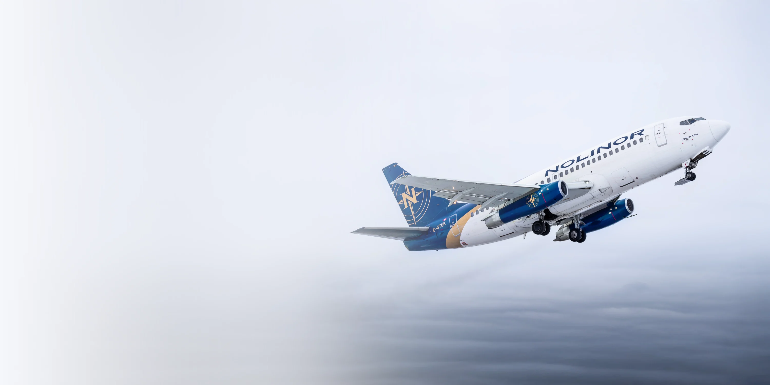 Un avion de la compagnie Nolinor décolle dans un ciel nuageux, montrant son design distinctif en bleu et blanc.
