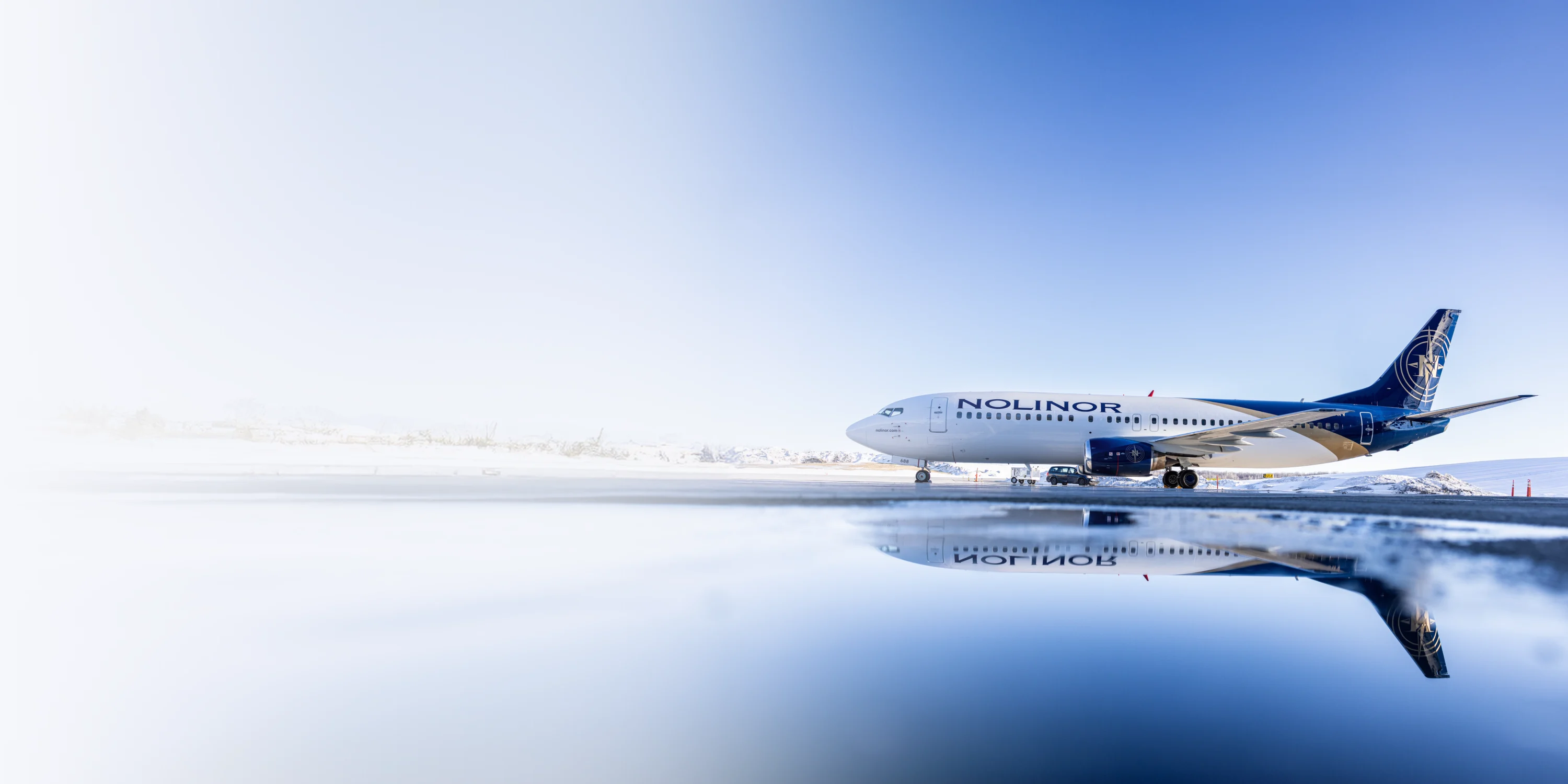 A Nolinor Aviation airplane is parked on a snowy runway, reflecting in a puddle, with a clear sky in the background.