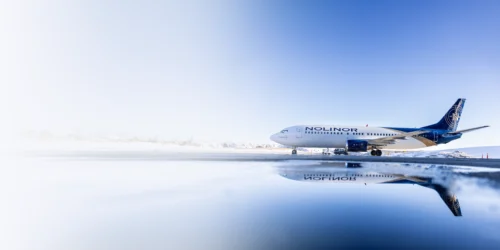 Un avion de la compagnie Nolinor est garé sur une piste, reflété dans une flaque d'eau, sous un ciel clair.