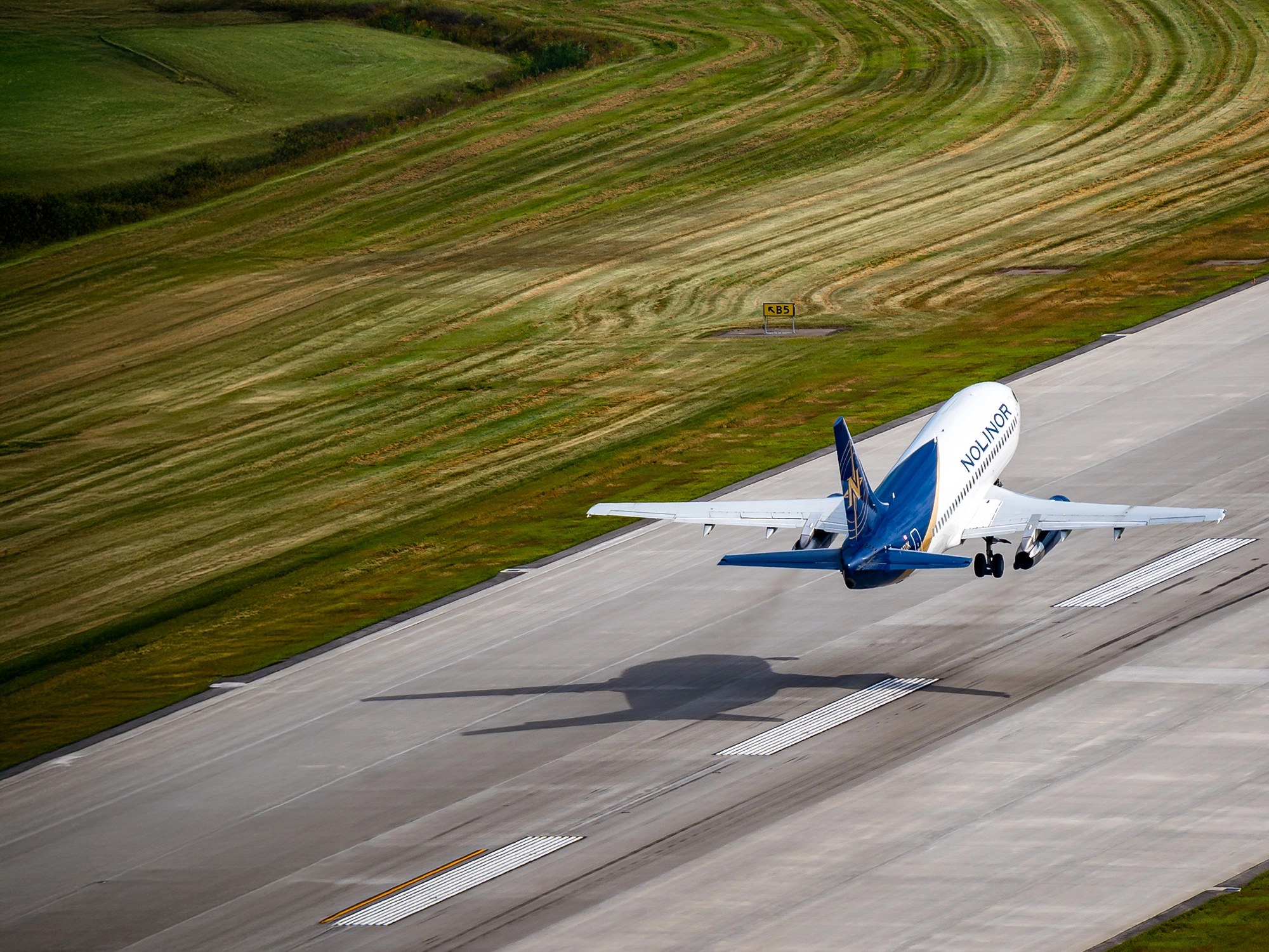Nolinor aircraft taking off from a runway, with its shadow visible on the ground and freshly mowed grass in the background.