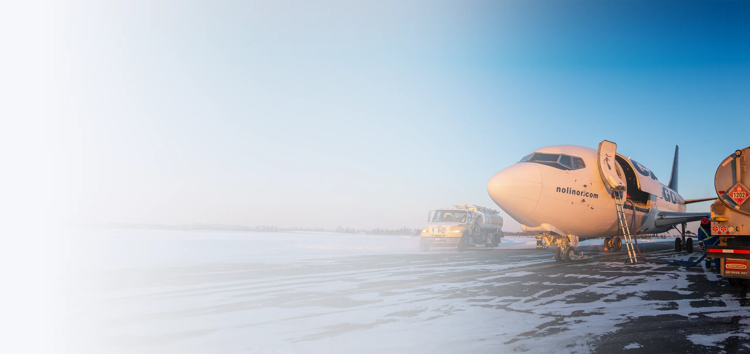 A plane is being refueled on a snowy runway, with a fuel truck parked nearby under a clear blue sky.