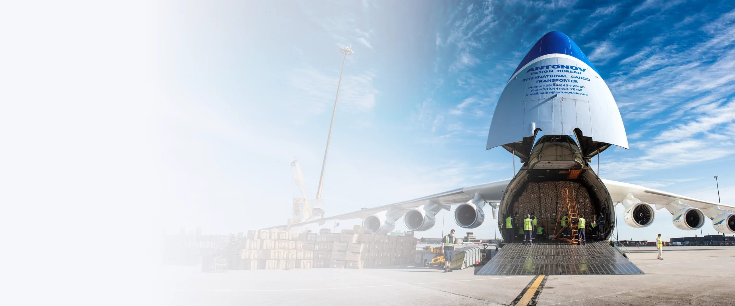 Antonov cargo plane with its nose raised, unloading cargo at a busy airport. Workers are visible inside the aircraft.